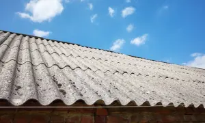Faliste płyty eternitowe, czyli jak wygląda azbest, pokrywają dach starego budynku. Pod nimi ceglana ściana, a nad nimi błękitne niebo z chmurami.