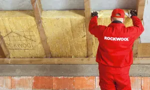 Pracownik w czerwonym stroju Rockwool montuje wełnę mineralną, wykonując ocieplenie murłaty od wewnątrz.